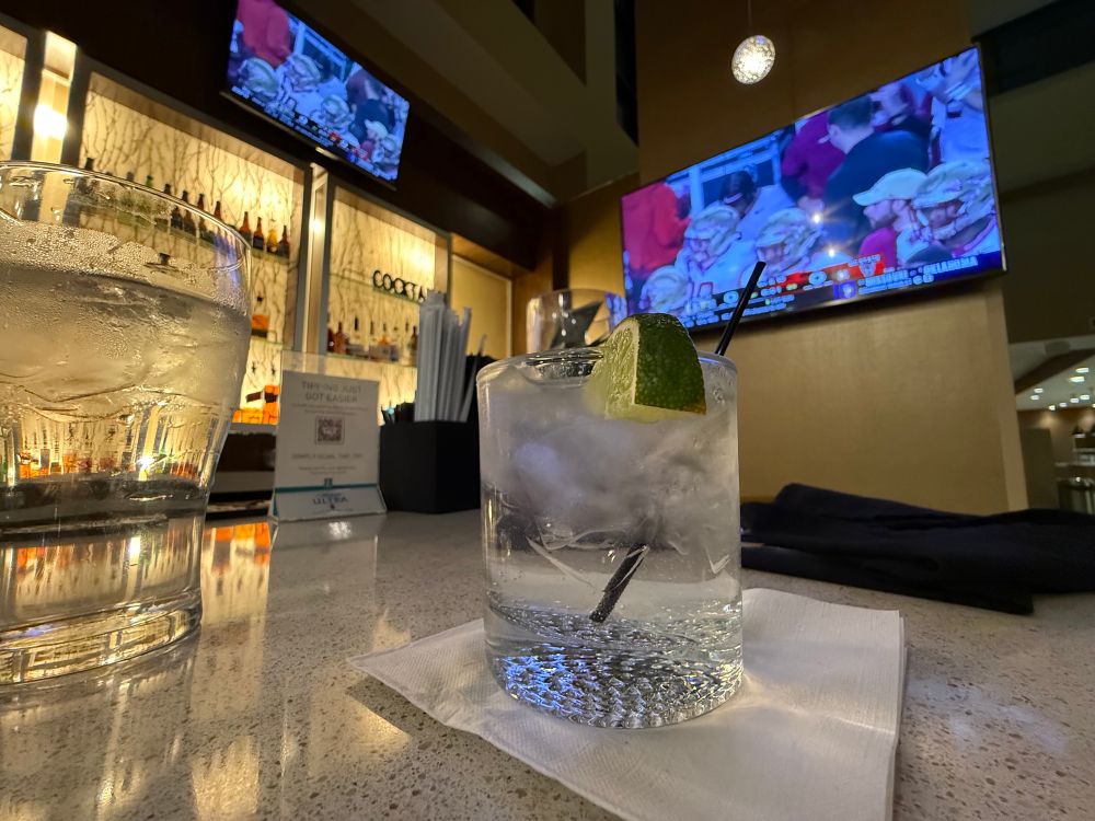 A rocks glass with a lime on the bar in this hotel. Florida State - NC State is on the TVs. In the glass is a gin and tonic with Drumshanbo Gunpowder Irish Gin