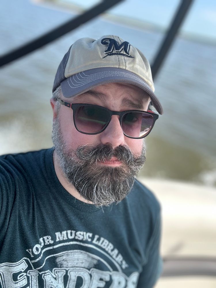 Selfie of the author, in sunglasses & a Brewers cap. Blurred in the background is the boat he is in and the water that boat is in.