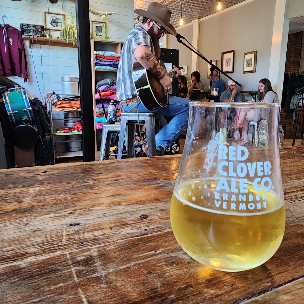 Red Clover Ale Company glass on a table with a man playing guitar in the background.