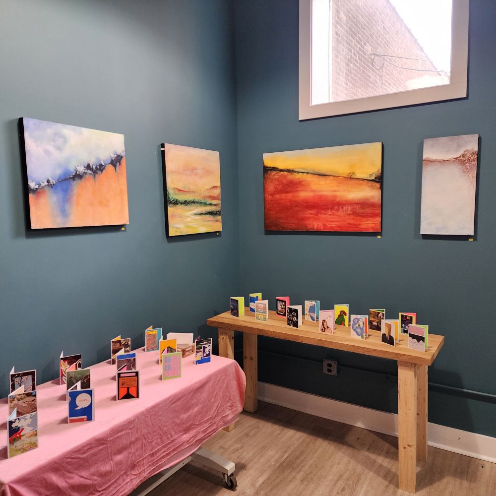 A small gallery with 4 abstract paintings hung in a blue/gray wall and two tables with artistic greeting cards.