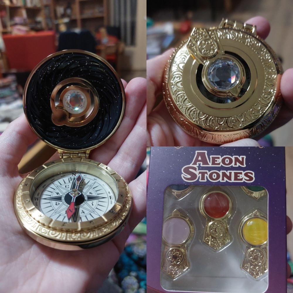Three pictures of a Wayfinder, a gold compass with a jewel set into the lid. One picture shows the compass needle and inside of the lid with a swirling night sky effect in enamel and glitter, one shows the closed wayfinder with a clear gemstone affixed to the top, and one shows a box labelled "Aeon Stones" with different coloured gemstones that can be swapped out with the one attached to the wayfinder.