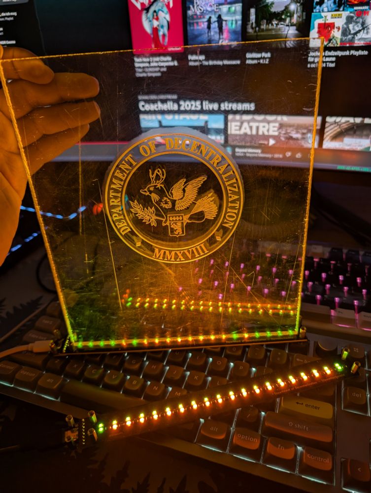 Holding a acrylic glass sign that has a "Department of Decentralization" logo engraved resting on two PCBs with green and orange LEDs illuminating the sign.