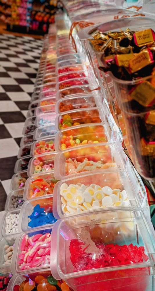 Eine Auslage von drei Reihen von durchsichtigen Plastikbehältern gefüllt mit Bonbons und bunten Süßigkeiten. Aus den Behältern füllt man die gewünschte Menge in die Tüte für die lördagsgodis. 