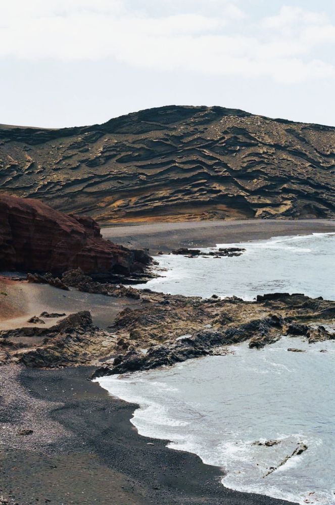 Küstenlandschaft mit schwarzem Sandstrand, felsigen Klippen und hügeligem Gelände im Hintergrund.