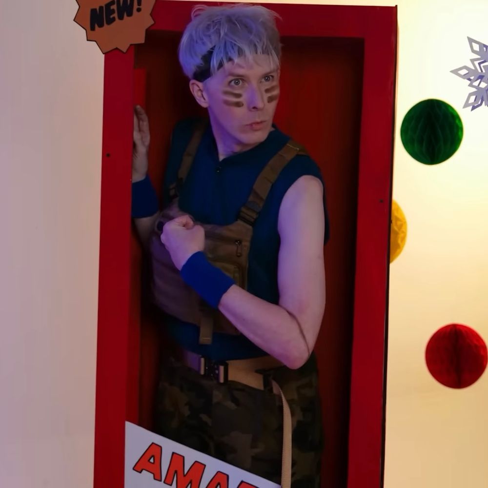 phil lester dressed as an action figure with war paint stripes on his cheeks. he has a bandana on his hair. he is wearing a blue muscle shirt and blue wrist warmers. he has camo trousers on and a brown chest pack on. he is pouting and flexing his bicep. he is standing in a life size red doll box