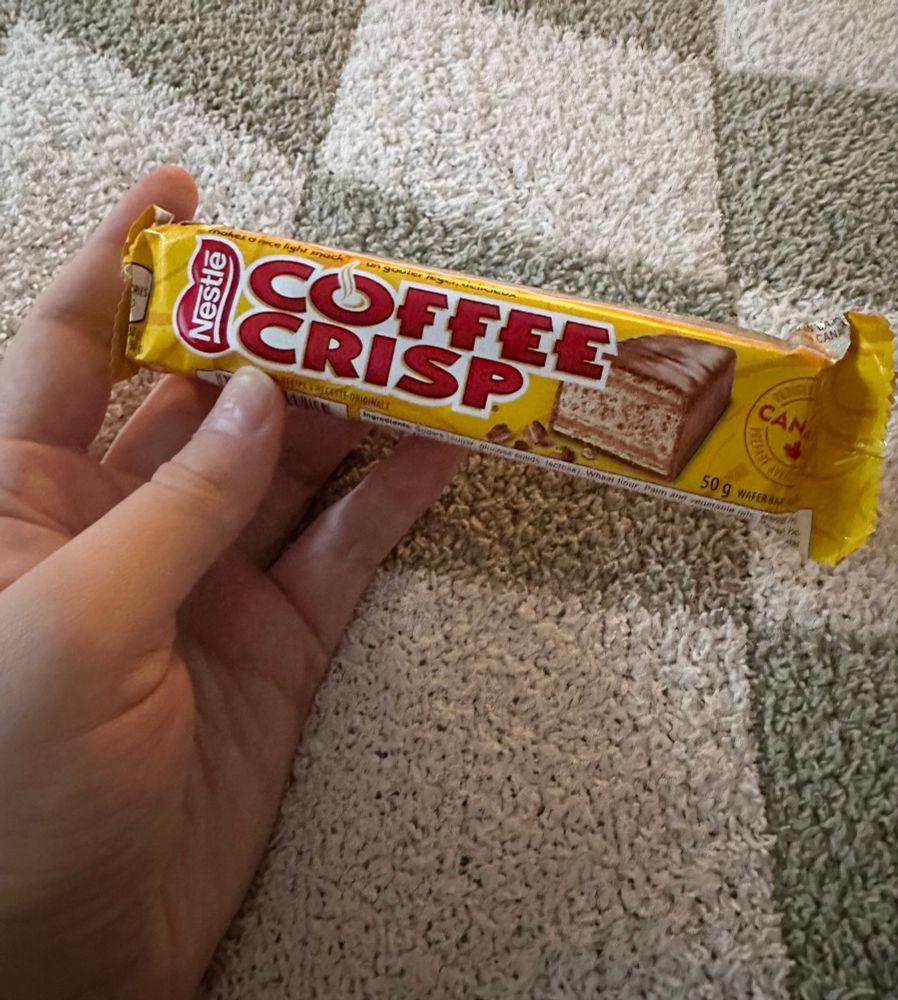 A Caucasian woman’s hand holds a yellow candy bar that reads “Coffee Crisp” in red block letters. 