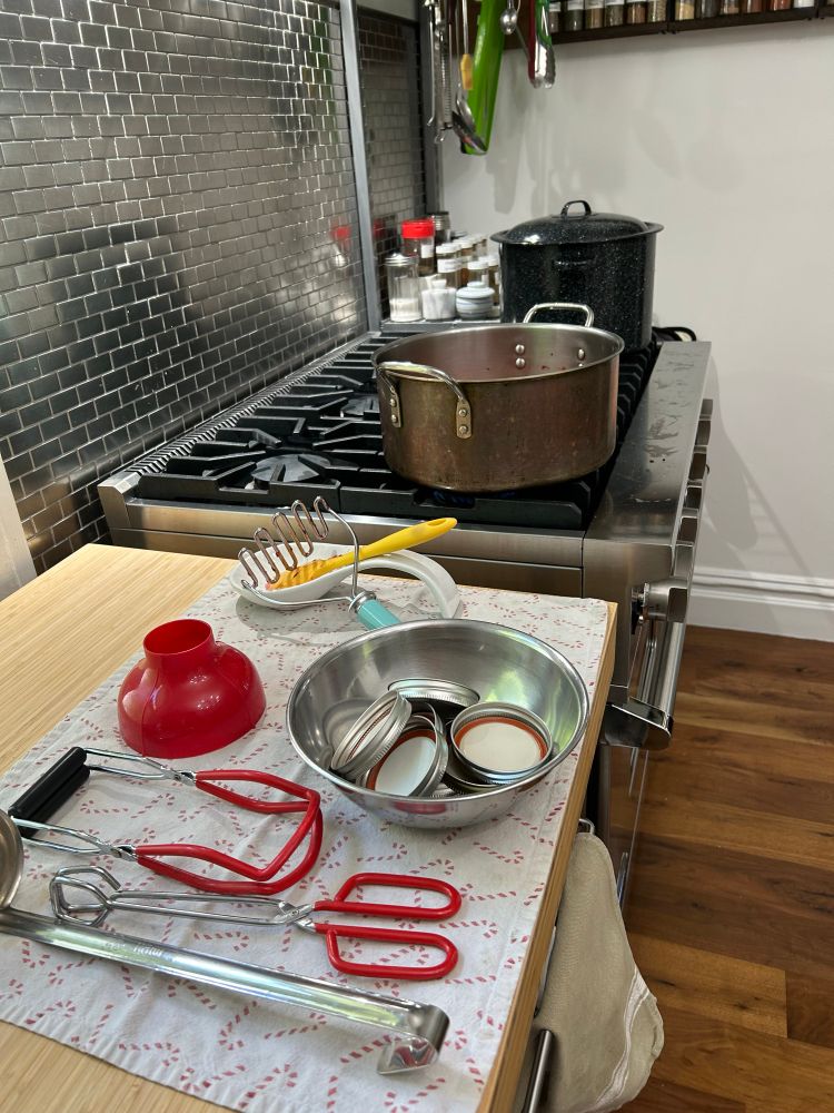 Array of jam making tools on a towel on a counter next to a stove with a large pot of jam and a canning kettle on it.
