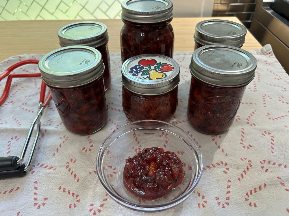 Six jars of marmalade and a little bowl of leftovers on a towel on a counter. Jar lifting tongs are next to them.