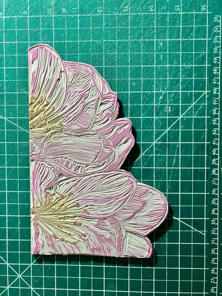 My carved Lino block shows two apple blossoms the size of salad plates with two flat edges that meet at a corner and the other edges tracing the petals. It’s sitting on a green cutting mat with half inch grid in white. The block is tinted from use. The petals are pinkish, the stamens pale green and the pollen rusty orange.