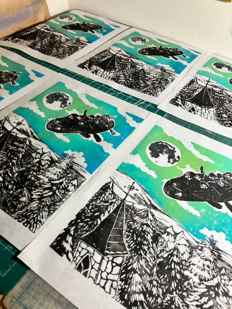 Several copies of my Lino block print shows a night sky with moon and clouds and flying canoe full of paddling voyageurs over rolling countryside with fields and trees covered in snow and a church steeple in the foreground. They are laid out in a grid on a tabletop to dry.