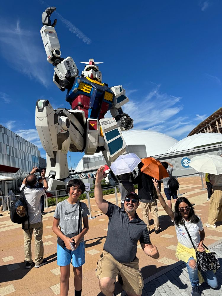 Squating in front of the rx-78-2 gundam on one knee with one hand raised in the air, mimicking the Gundam’s pose. On both sides James is flanked by his son and daughter, the later mimicking the same pose while the son smiles on with a deck of Pokemon cards in his hand.