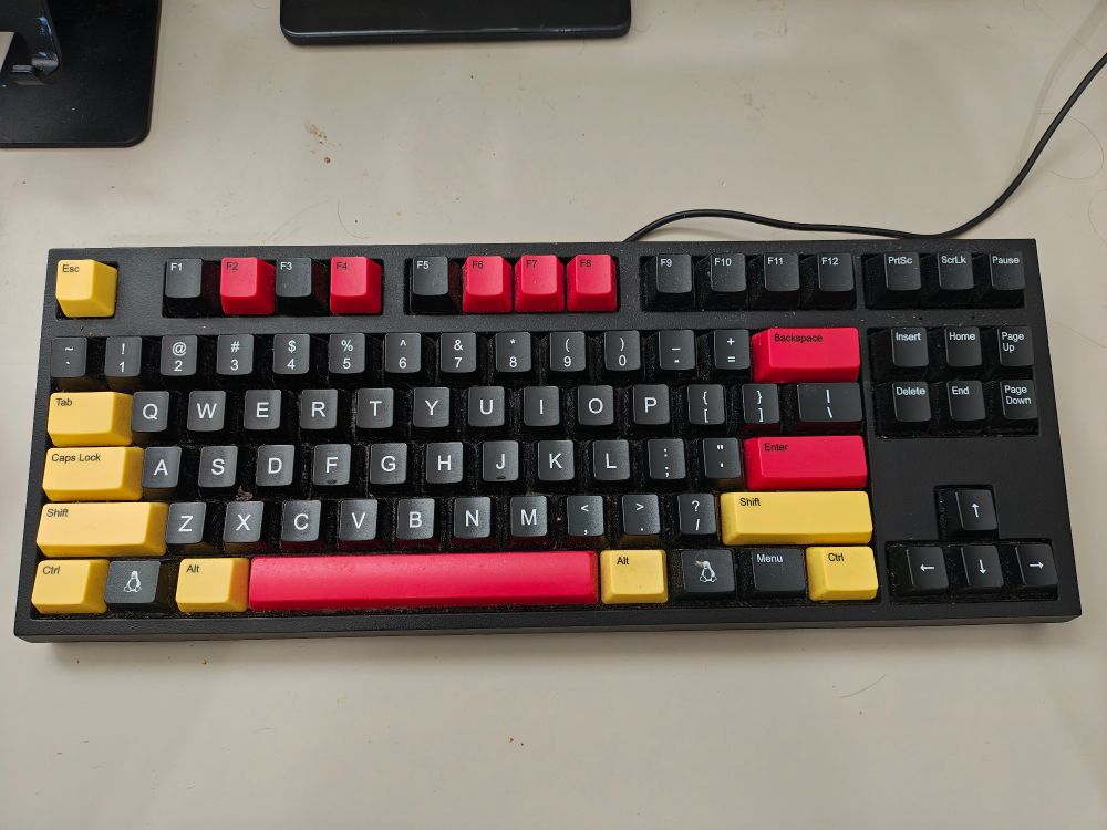 Meu teclado mecânico, preto com as teclas de controle (tab, caps, ctrl, shift esc) em amarelo; backspace, space e enter em vermelho, assim como as teclas F2, F4, F6, F7 e F8.
