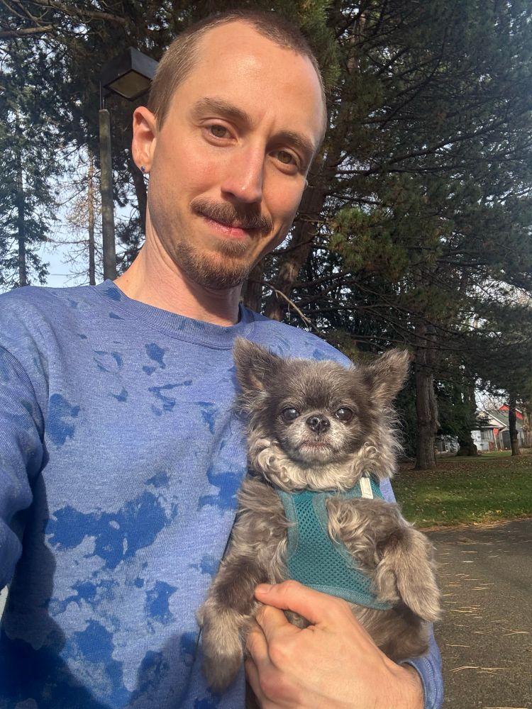 I’m holding up Jem, a long-haired chihuahua with grey fur. I’m wearing a blue sweater.
