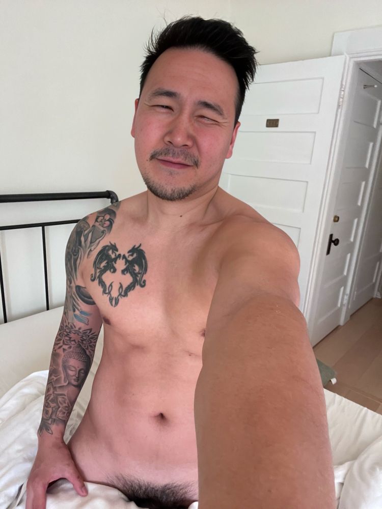Naked selfie of man in bed with pillow covering legs showing visible pube