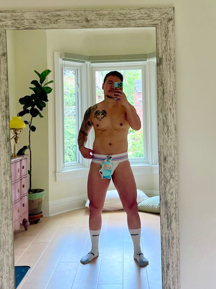 Mirror selfie of man wearing nothing but jockstraps and white socks with a labubu hanging over crotch