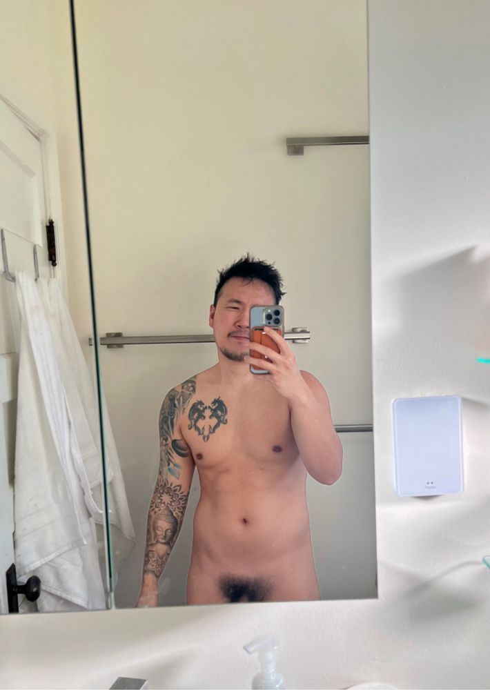 Man posing naked in front of mirror showing base of penis and bushy pubes