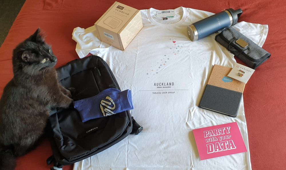 Tableau swag pack spread on a red surface, displaying: a backpack, a pair of socks, a greetings card saying "party with your data", a notebook with the Salesforce branding, a electronics organiser, speakers, a water tumblers and a custom-made tshirt with the winning design for the Auckland TUG tshirt design contest, with the print designed by T. from Data Rocks. The print sows a map of New Zealand using the Tableau logo as a pattern, with a Heart placed where Auckland is located. The text underneath the illustration reads "Auckland Tamaki Makaurau, Tableau User Group". The swag pack is being appreciated by a black cat, Ozzy, Data Rocks Chief Purring Officer.