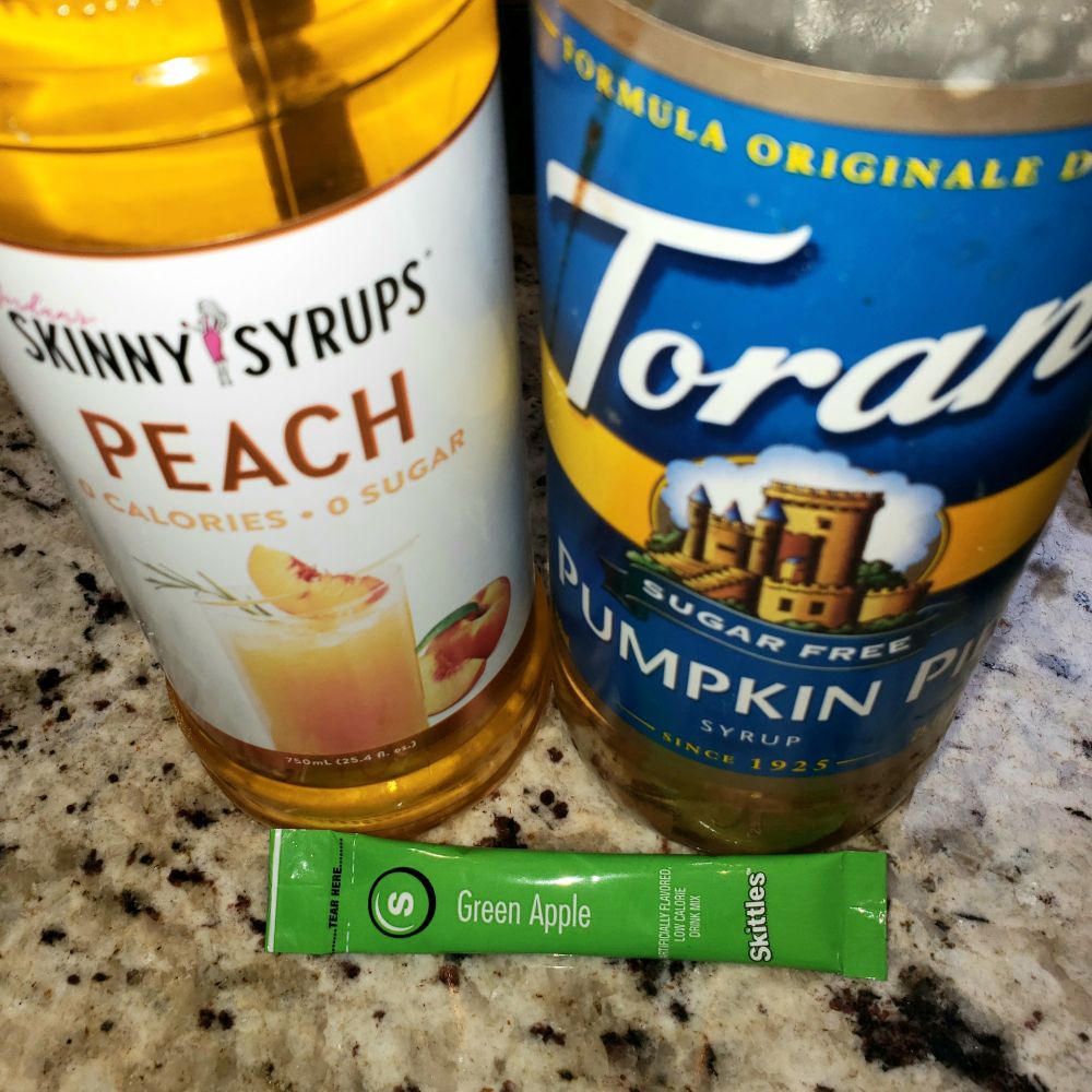 A packet of Skittles-brand Green Apple drink mix, a bottle of Skinny Syrups-brand Peach syrup, and a bottle of Torani-brand Sugar-Free Pumpkin Pie syrup.