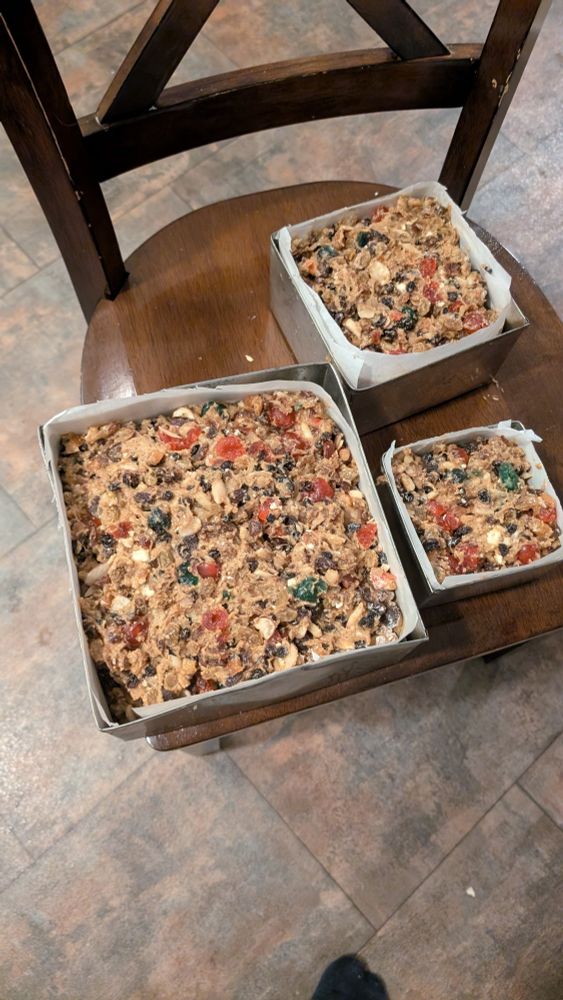 Three square pans full of unbaked dark fruitcake, one large,one medium, and one small are sitting on a brown kitchen chair.