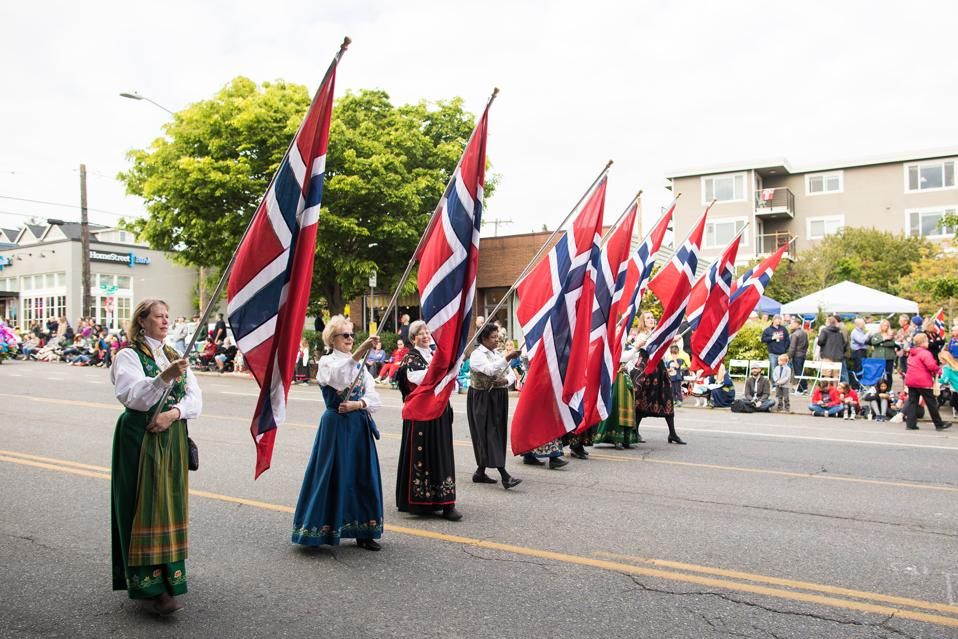 A picture of the Norwegian Constitution Day parade in Seattle. A row of people are walking down the street wearing traditional Norwegian costumes and holding large Norwegian flags.