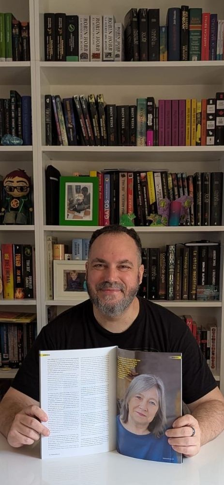 Traductor sonriente posando delante de una estantería llena de libros con la revista Windumanoth abierta por la entrevista a Robin Hobb que contiene.