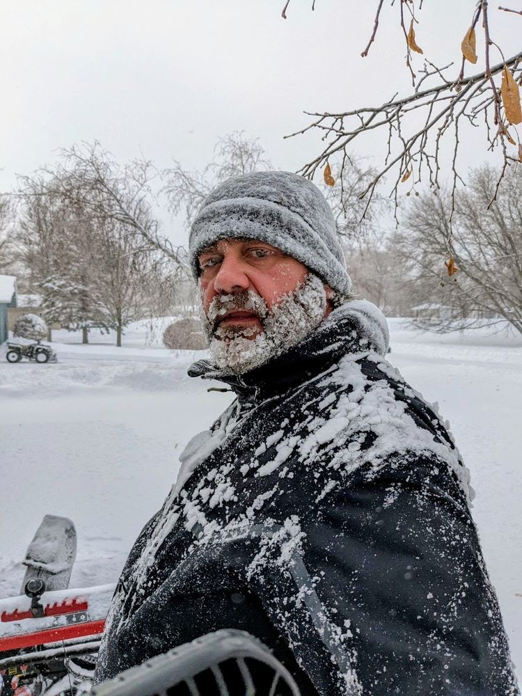 Photo of an old white guy, standing outside in the winter, next to a snowblower, wearing a winter hat and coat, completely covered with snow, his beard caked with ice, looking like some kind of wampa on the ice planet Hoth.