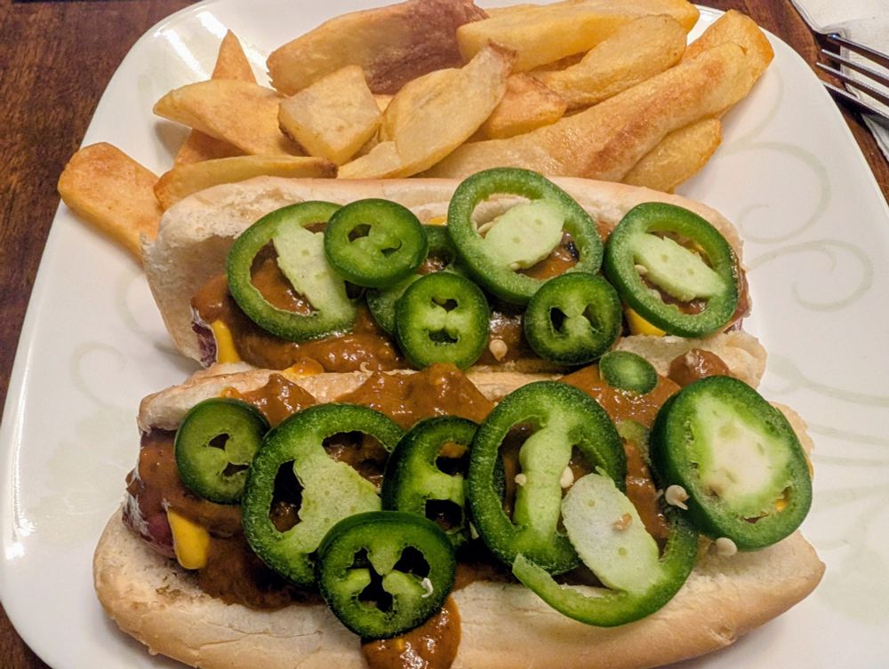 Photo of a dinner plate loaded with steak fries, and two grilled Nathan's Famous jumbo beef franks, on hot dog buns, topped with yellow mustard, no bean chili, and fresh jalapeño slices.