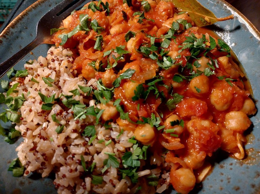 A serving of Greek-style braised chickpeas with tomatoes and orange lies to the right of a brown-speckled and brown-edged blue ceramic bowl. Rice with quinoa lies to its left. The dish is topped with roughly chopped flat leaf parsley.