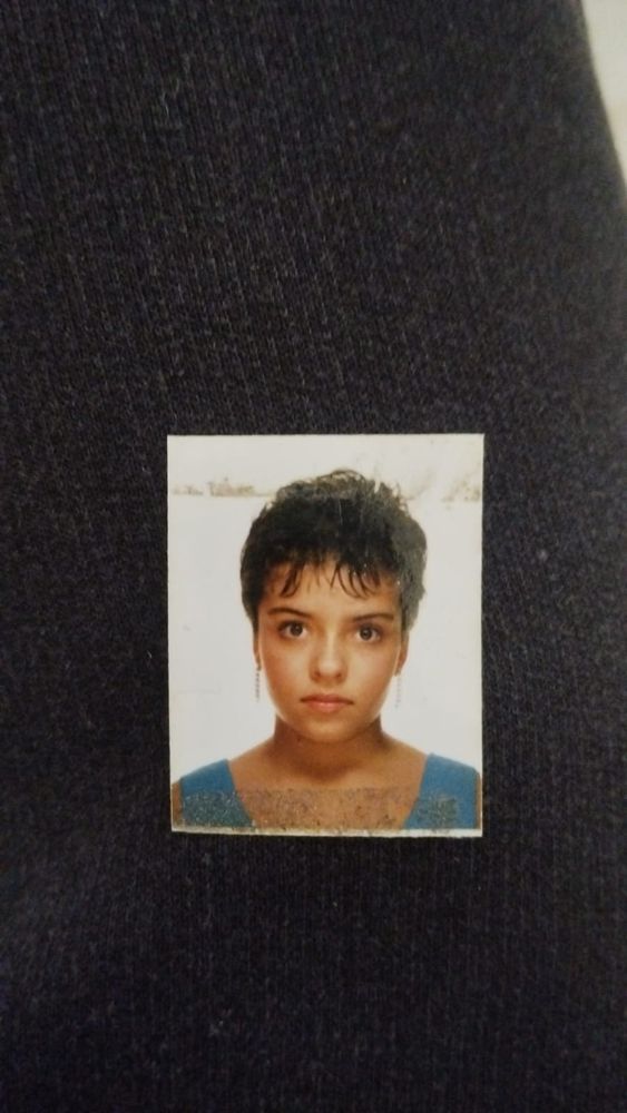 A imagem é uma fotografia em formato vertical, semelhante a uma foto de passaporte ou identidade, com foco no rosto de uma criança ou adolescente.
Descrição da Fotografia:
 * Pessoa: O rosto está centrado e enquadrado. A pessoa tem cabelo curto, escuro e encaracolado, com caracóis definidos em torno da cabeça. A testa é visível sob os cachos.
 * Feições: Os olhos são grandes e de cor castanha. A boca está fechada numa expressão séria ou neutra. A pessoa usa brincos pequenos e pendurados (argolas finas ou dangles).
 * Roupa: A parte superior da roupa visível é azul-clara (possivelmente um top ou alça de camisola).
 * Formato e Fundo: A fotografia é um pequeno retângulo (tamanho de foto 3x4) com um fundo branco (fundo de estúdio fotográfico), colada sobre um tecido ou superfície escura e texturizada, que ocupa a maior parte da imagem. A pequena foto parece estar ligeiramente gasta nas bordas, indicando a sua idade.
