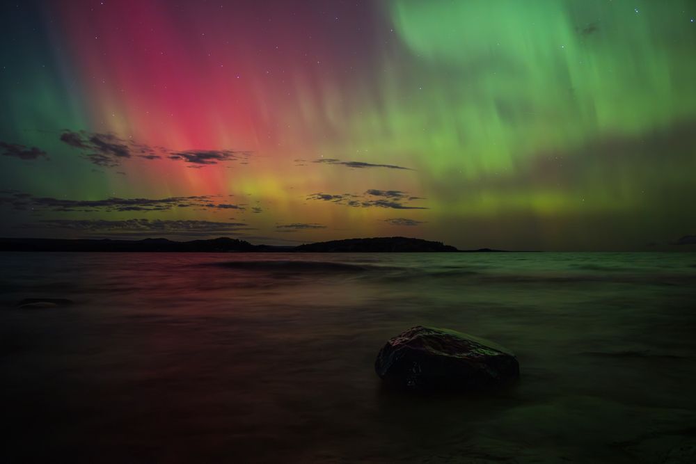 Aurora borealis over Lake Superior during the overnight period between Oct. 7 and Oct. 8, 2024, near Marquette, MI. Greens and pinks/reds are the theme in this photo, with a single rock serving as the driving foreground element.