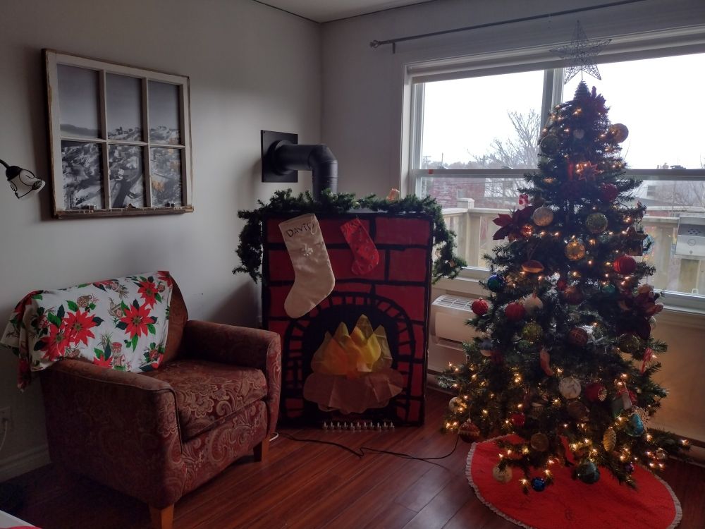 A cozy space with a cardboard cut out fireplace in the middle with two stockings, a Christmas tree to the right, and on the left a arm chair with a nice flowered throw over it. It just looks super homey and inviting.

