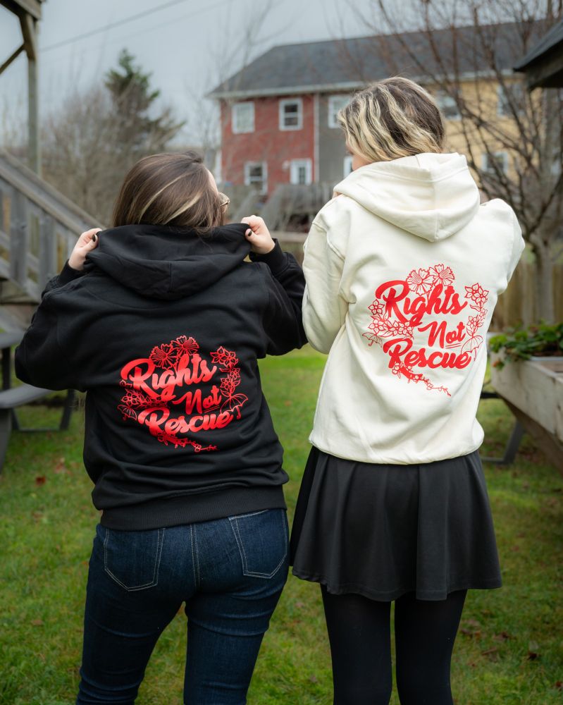 Two people standing outside on a chllly looking day. They are facing away from the camera and the person on the left is wearing a black hoodie and the eprson on the right is wearing an ivory hoodie. The hoodies say "Rights Not Rescue" and the words are surrounded by flowers and a red umbrella.