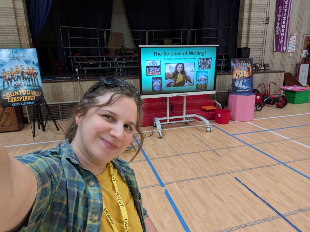 Selfie of me, a white person with brown hair pulled into a ponytail and sunglasses on my head, wearing a yellow shirt with an open green button up shirt over it, standing in a gymnasium in front of a projector showing my school visit slideshow and some signs with my book covers.