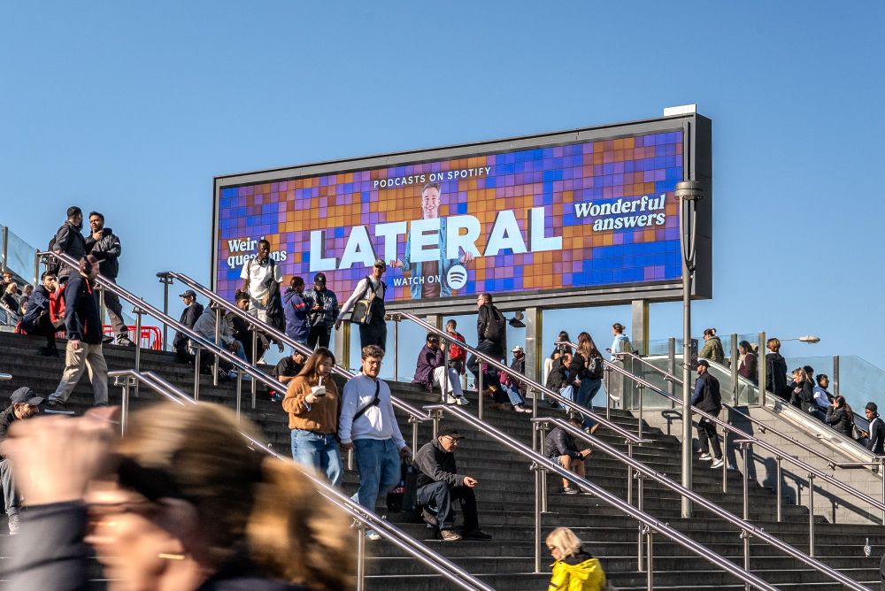 An electronic billboard for 'Lateral with Tom Scott' in front of a large set of steps with people walking up and down them.