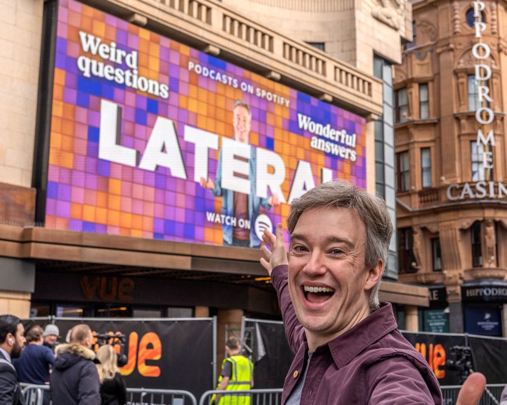 Tom Scott gesturing towards an electronic billboard for 'Lateral with Tom Scott' in Leicester Square, London