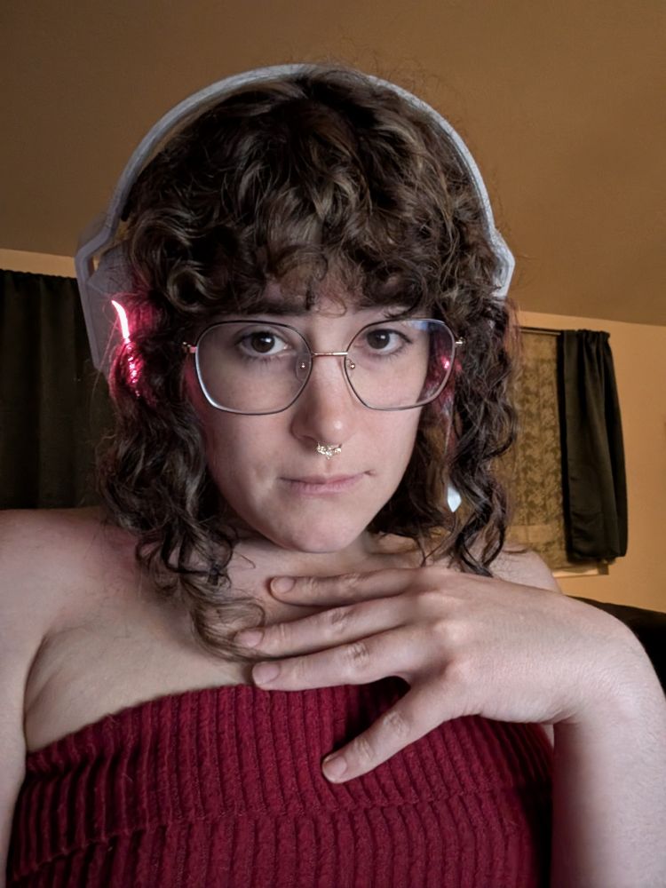 Trans girl with brown hair and a red tank top taking a selfie with a nose ring and glasses. Her hand is up towards her chest covering it slightly 