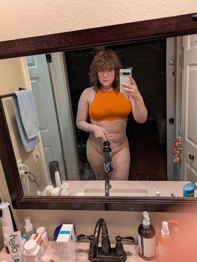 Trans girl taking a mirror selfie with an orange top covering her crotch with a water bottle 
