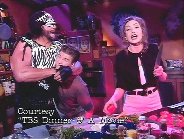 A shot from "TBS Dinner & A Movie", where guest "Macho Man" Randy Savage has host Paul Gilmartin in a headlock while co-host Annabelle Gurwitch addresses the camera, knife and tomato in hand 