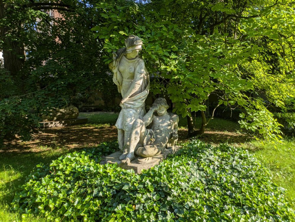A wide shot of a statue in a lush greed garden, the statue depicts a man laying on the floor with his hand on his brow, and a woman walking beside him moving her clothes as to not hit him on the way past. The statue was built in 1883. 