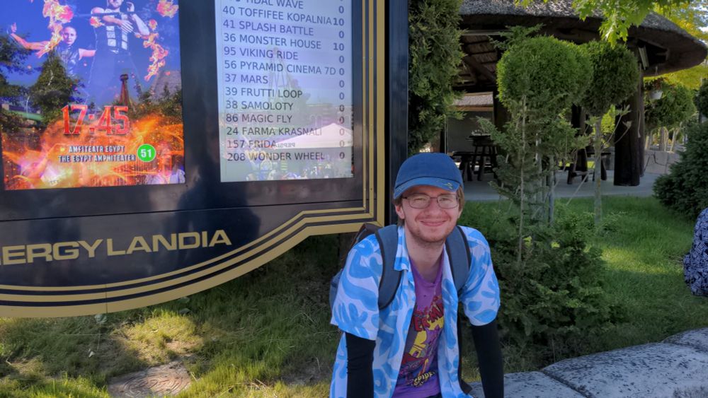 Keenan sat on the side of a path in a theme park, wearing a bright blue and white shirt with a Spider-Man undershirt, a blue cap. Enjoying the break.