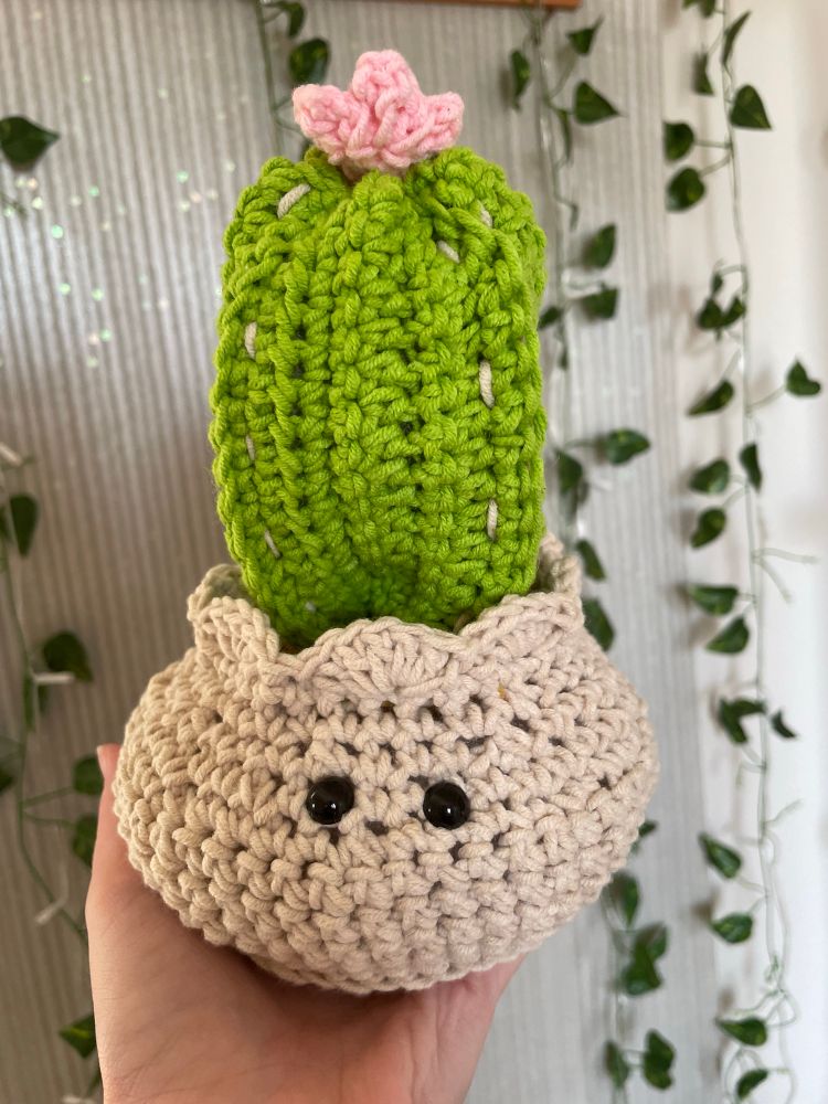 fencepost cactus in a pot that i have crocheted. lil round white pot with cute eyes, green cactus bit with some white needles and a pink flower on top 