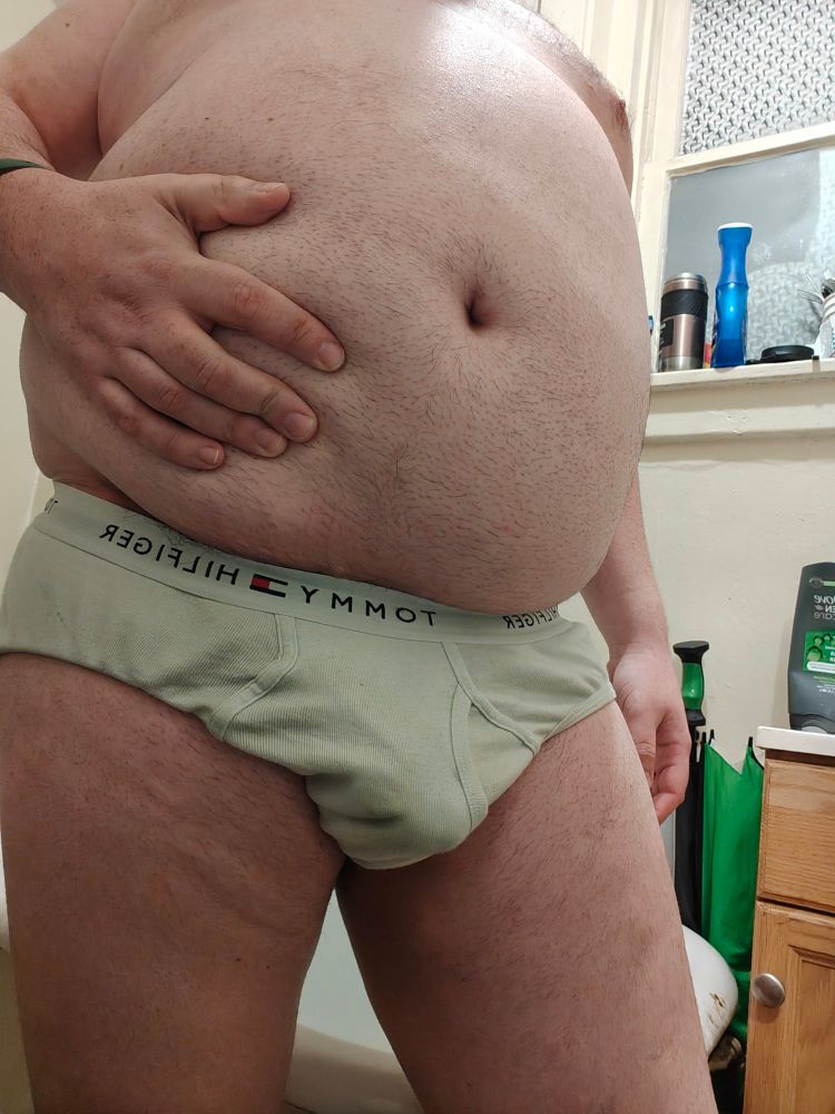 A white, fat man wearing white Tommy Hilfiger briefs squeezing his belly and posing for the camera.