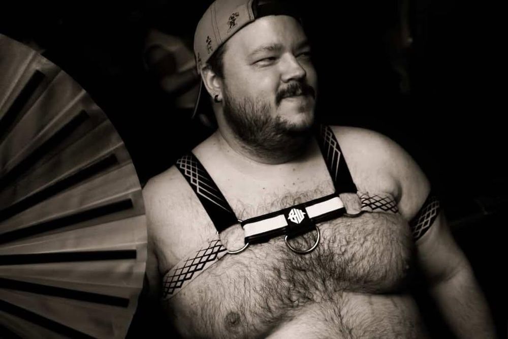 Black and white photo of a chubby man, shirtless and in a harness while holding a fan
