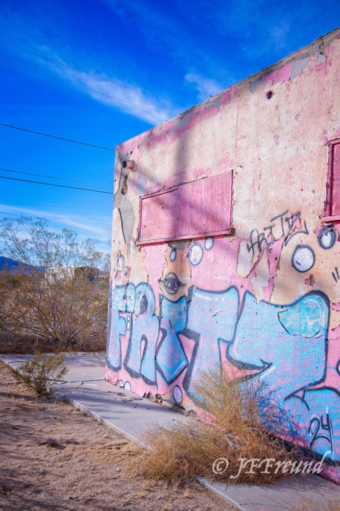 Graffiti FRiTZ in blue on plaster peach colored plaster wall.  Shadow of a Telephone pole forms a cross.  Weeds grow through sidewalk.  Blue sky in the background with high white clouds.  The building is clearly abandoned