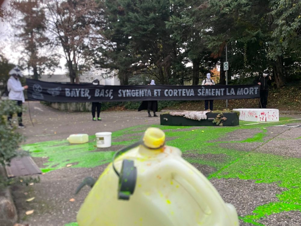 Une banderole XR avec écrit "Bayer Syngenta et Corteva sèment la mort". On voit un bidon jaune (?) au premier plan et une tâche verte avec des bidons renversés sur le sol.