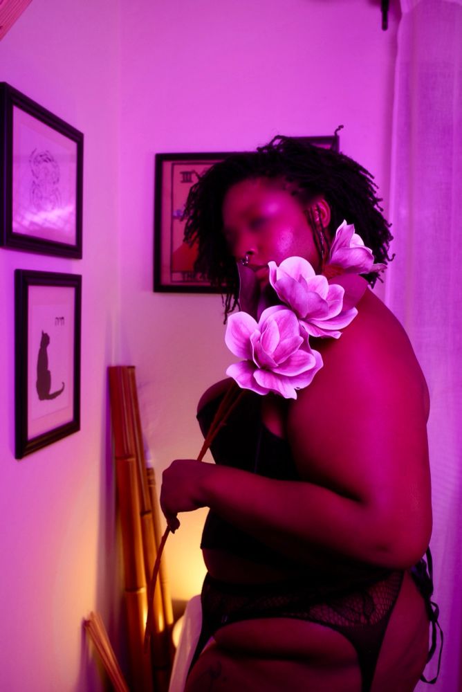 Sage, bathed in purple light, holds a flower over their shoulder. 