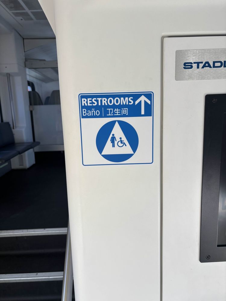 A new onboard sign inside a Caltrain EMU  indicating the direction to the restrooms. The blue and white sign reads “RESTROOMS” with translations in Spanish and Chinese, and includes an upwards arrow. Below the text is an accessibility symbol featuring a person and a wheelchair icon. 