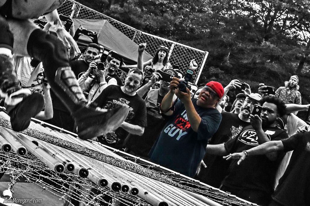 greyscale photo of a wrestling crowd gathered around a barbed wire trampoline. Up against it, the solo subject in color is Lyle Williams holding his camera aimed at the cropped out action.