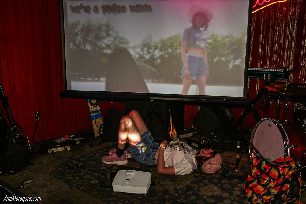 Trust Fund Ozu lies down in front of a screen which has a video of herself playing