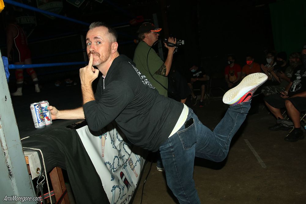 Dan Barry leans on a wrestling ring while holding two beers while kicking a leg back while facing the camera with a finger over his lips
behind him is a camera operator and wrestling crowd